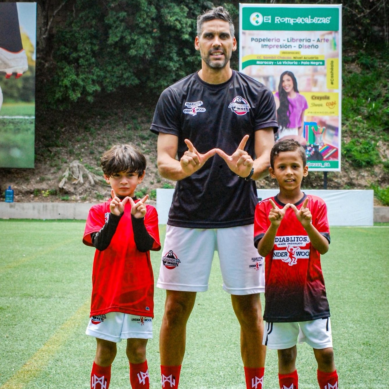 Entrenador con dos niños en campo de fútbol, todos haciendo un gesto de "W".